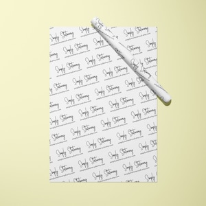May include: White wrapping paper with a repeating pattern of the text "Simply Stationery" in black cursive writing. A white pen is rolled up on top of the paper.