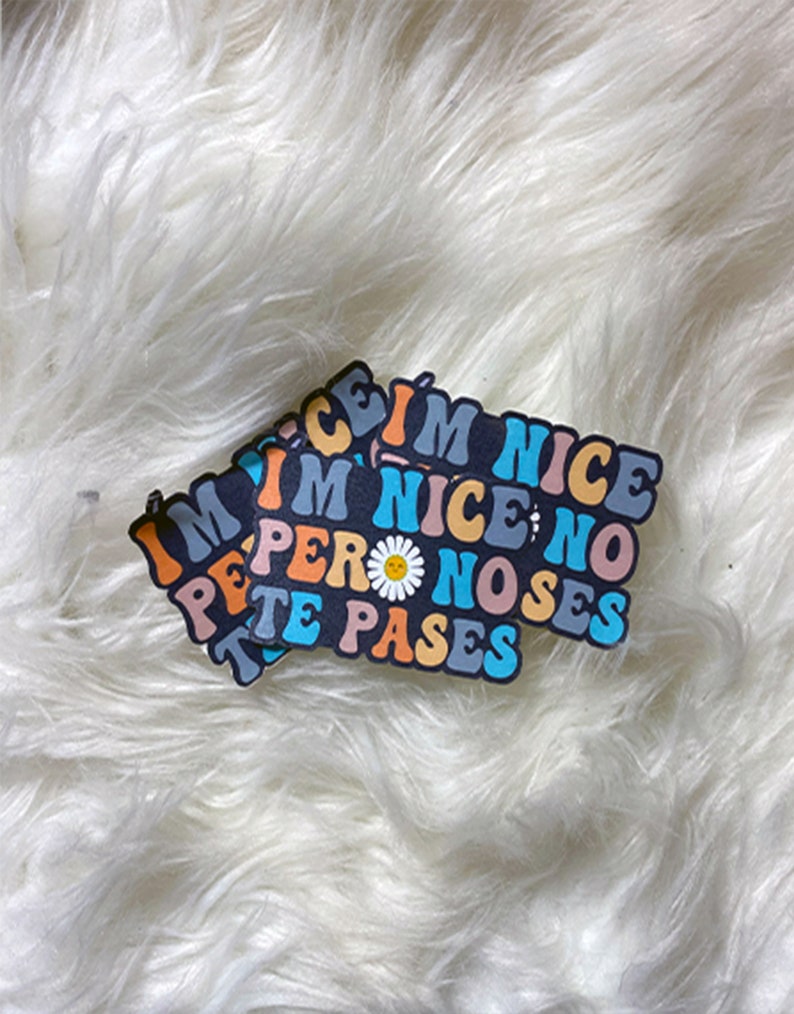 May include: A sticker with the text "I'm nice; no pero noses te pases" in a rainbow color scheme with a daisy in the middle. The sticker is on a white fluffy surface.