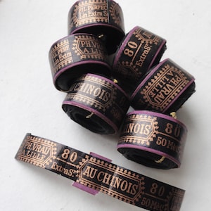 Könnte beinhalten: Eine Sammlung schwarzer Garnrollen mit goldfarbenen Schriftzügen und lila Akzenten. Die Rollen tragen Aufschriften wie "AU CHINOIS" und "80 Extras". Das Garn ist um die Rollen gewickelt, eine Rolle ist abgerollt. Jede Rolle enthält ca. 50 Meter Garn.