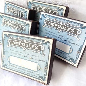 Könnte beinhalten: Eine Sammlung von Vintage-Ampullenboxen. Jede Box hat ein hellblaues Etikett mit dem Wort "AMPOULES" in einer dekorativen Schriftart. Die Boxen haben dunkelbraune Seiten und sind auf einer weißen Oberfläche angeordnet.