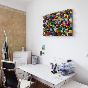 Op de afbeelding: Een wit bureau met een witte stoel en een witte commode in een thuiskantoor. Het bureau heeft verschillende kantoorbenodigdheden, waaronder pennen, potloden, een computermuis en een stapel doorzichtige plastic bakken. Boven het bureau hangt een kleurrijk geometrisch wandkunstwerk.