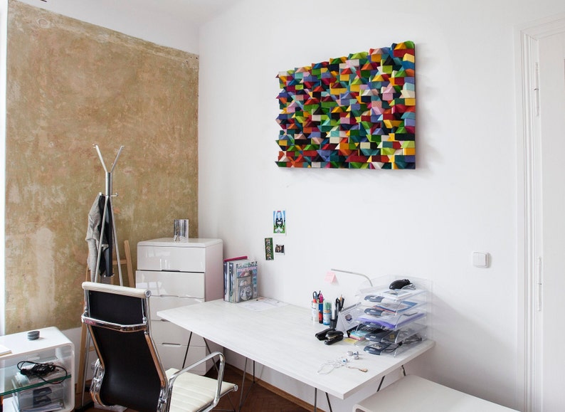 May include: A white desk with a white chair and a white dresser in a home office. The desk has a variety of office supplies on it, including pens, pencils, a computer mouse, and a stack of clear plastic trays. There is a colourful geometric wall art piece above the desk.