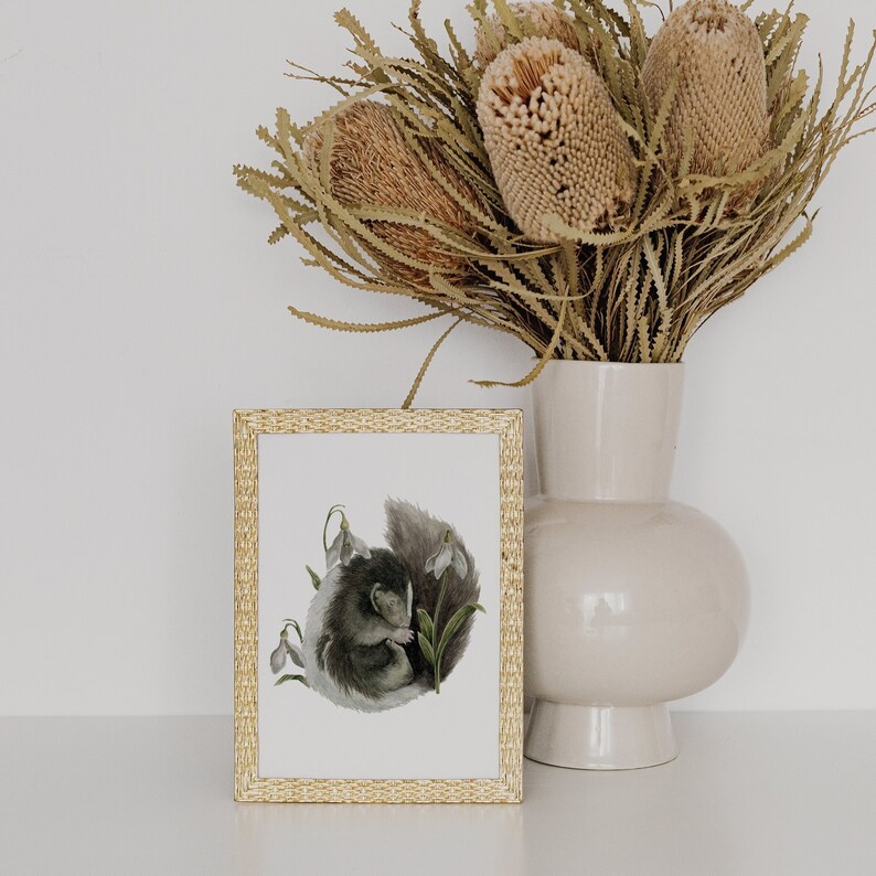 May include: A framed illustration of a small animal curled up among white snowdrop flowers. The frame is gold with a woven pattern. The background is a white wall with a vase of dried flowers.