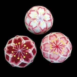 Puede incluir: Tres bolas Temari hechas a mano con intrincados diseños florales. Las bolas presentan patrones de hilo blanco, rojo y rosa, con detalles amarillos en el centro de las flores. Las bolas Temari se muestran sobre un fondo negro.