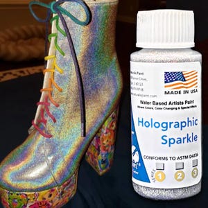 May include: A holographic, iridescent ankle boot with rainbow laces and a platform heel. Next to it is a bottle of "Holographic Sparkle" water-based artist's paint, made in the USA.