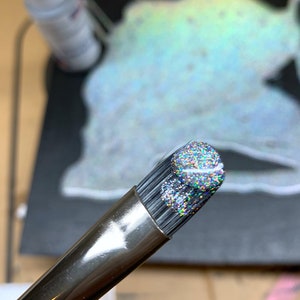 May include: Close-up of a makeup brush with a silver handle and a head covered in iridescent glitter. The bristles are black. The background is blurred, with hints of other objects.