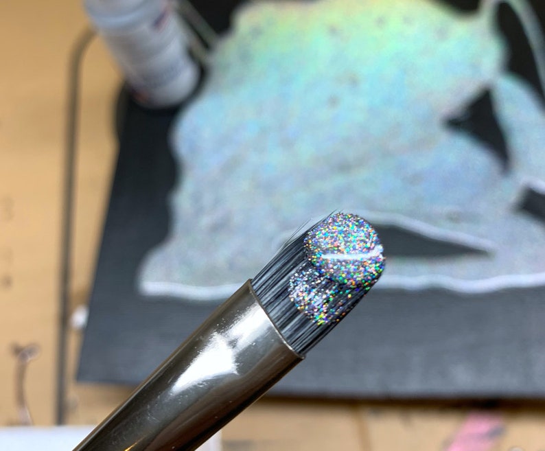 May include: Close-up of a makeup brush with a silver handle and a head covered in iridescent glitter. The bristles are black. The background is blurred, with hints of other objects.