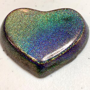 May include: A heart-shaped object with a shimmering, iridescent finish. The surface displays a blend of colors, including green, blue, and purple, with a speckled texture. The edges of the heart are rounded and smooth.
