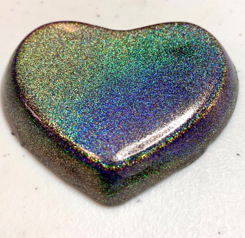 May include: A heart-shaped object with a shimmering, iridescent finish. The surface displays a blend of colors, including green, blue, and purple, with a speckled texture. The edges of the heart are rounded and smooth.