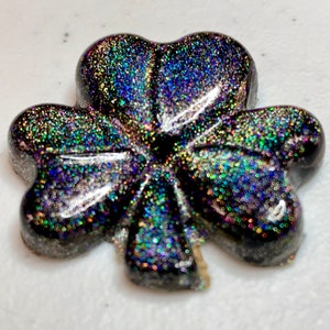 May include: A glittery, black shamrock-shaped resin charm. The charm is covered in iridescent glitter that reflects multiple colors. The shamrock has three rounded leaves and a heart-shaped center. The charm is on a white surface.