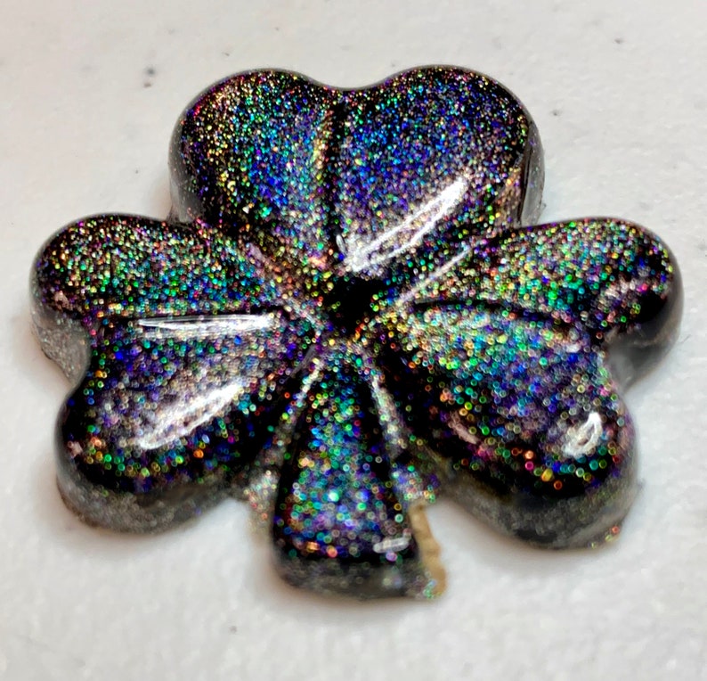 May include: A glittery, black shamrock-shaped resin charm. The charm is covered in iridescent glitter that reflects multiple colors. The shamrock has three rounded leaves and a heart-shaped center. The charm is on a white surface.
