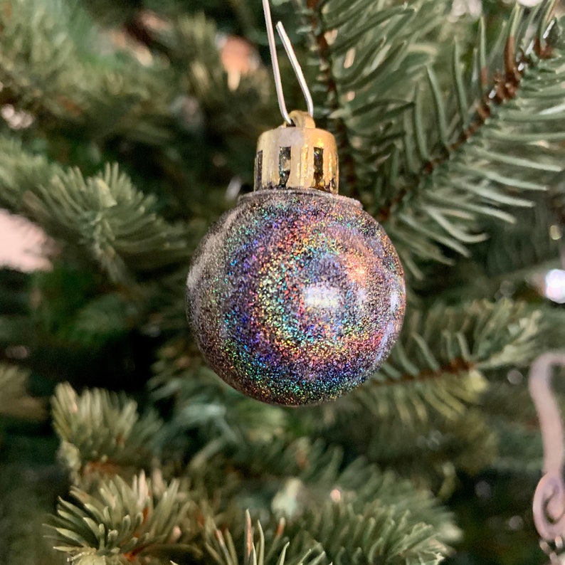 May include: A close-up of a Christmas tree ornament. The round ornament is covered in iridescent glitter, reflecting multiple colors. It hangs from a silver wire and has a gold-colored metal cap. The background is a green Christmas tree.