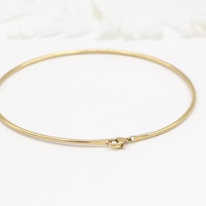 May include: A fine gold-coloured necklace with a minimalist design. The necklace is a thin, circular band that fastens with a small clasp. The jewellery is likely made of gold or a gold-coloured metal. The necklace is a classic piece.