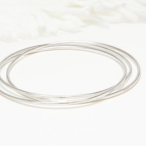 May include: Three silver bangle bracelets stacked on top of each other on a white surface.