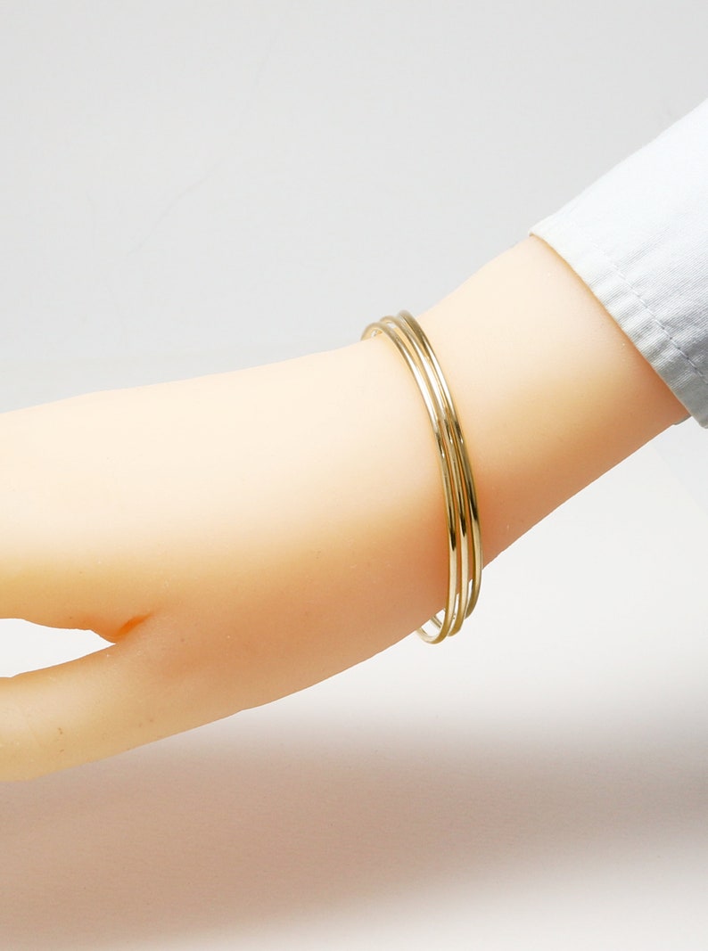 K&ouml;nnte beinhalten: Drei goldene Armreifen, die auf einem Handgelenk &uuml;bereinander liegen. Die Armreifen sind schlicht und haben eine polierte Oberfl&auml;che.