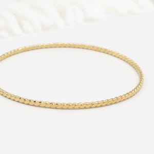 May include: A gold-colored bangle bracelet with a textured, twisted rope design. The bracelet is circular and sits on a white surface. The jewelry is a simple, elegant accessory.