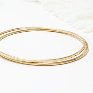 Puede incluir: Dos pulseras de oro, una ligeramente más grande que la otra, sobre una superficie blanca.