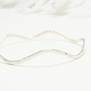 May include: A silver-colored, hammered-finish bangle bracelet with a wavy design. The bracelet has a simple, elegant style and is likely made of sterling silver or a similar metal. The bracelet is shown on a white background.