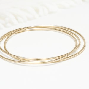 May include: Three gold bangle bracelets stacked on top of each other on a white surface.