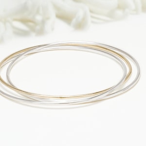 Könnte beinhalten: Drei Armreifen, ein goldener und zwei silberne, übereinander auf einer weißen Oberfläche.