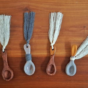 May include: Four decorative spoons with tassels, arranged on a wooden surface. The spoons are made of wood and ceramic, with varying colors and tassel hues. The tassels are white and gray.