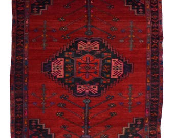 May include: A rectangular area rug featuring a deep red background with a large central diamond medallion in black, blue, and pink. The rug displays symmetrical tree-like motifs and geometric patterns throughout, finished with a short fringe edge.