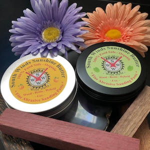 May include: Two jars of South Winds Sunshine Butter wood-resin hybrid sanding paste. The jars are labeled with the product name, ingredients, and size. The jars are surrounded by wooden blocks of various colors and grains.