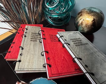 May include: Three small, bound journals with black rings. The covers are made of wood and are painted in different colors: gray, red, and brown. The journals have inspirational quotes printed on the covers.  The journals are stacked on top of each other.