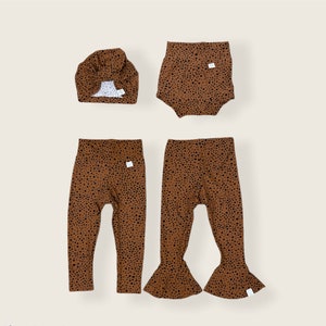 May include: A set of brown and black patterned clothing for a baby or toddler. The set includes a hat, a pair of leggings, and a pair of bloomers.