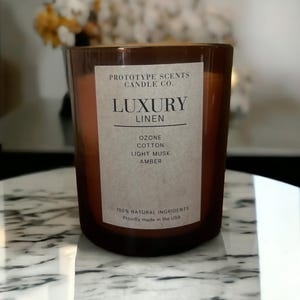 May include: A brown glass candle jar with a white label. The label reads "LUXURY LINEN" and lists scent notes. The candle is made with natural ingredients and proudly made in the USA.