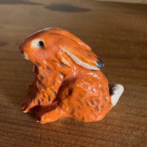 May include: A ceramic rabbit figurine in a sitting position. The rabbit is predominantly orange with a textured finish, black eyes, and a white tail. The ears have black tips. It rests on a wooden surface.
