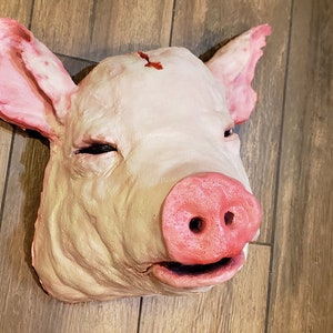 May include: A realistic pig head prop, made of a pink and beige material. The pig's snout is pink and has a small black nostril. The pig's eyes are closed, and the head is slightly tilted to the side.