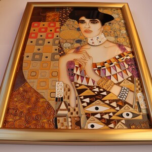 May include: A framed stained glass artwork depicting a woman with dark hair and a gold necklace. The woman is wearing a colorful dress with geometric patterns and gold jewelry. The background is a mosaic of gold, brown, and white.