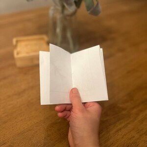 May include: A white blank booklet with a soft cover is open and being held by a hand. The booklet is on a wooden table.
