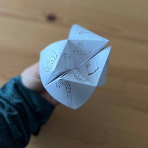 May include: A white paper fortune teller with black line drawings of a candy cane, a star, a Christmas tree, a stocking, a snowman, and a person wearing a robe.