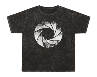 Może przedstawiać: Czarny T-shirt z białym nadrukiem obiektywu aparatu. Nadruk ma postarzony wygląd, jakby został wydrukowany na ceglanej ścianie.