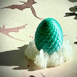 May include: A teal-colored dragon egg with a textured surface, resting on a bed of clear, crystal-like structures. The egg is the focal point, set against a backdrop of watercolor art featuring dragon silhouettes and foliage.