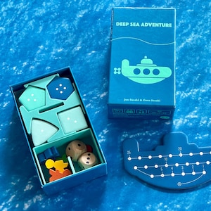 May include: A board game titled "Deep Sea Adventure" with a blue submarine theme. The game includes a box, game pieces, and a submarine-shaped game board. The box features a submarine illustration and the game's title.