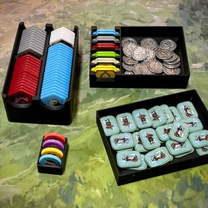 May include: A set of black plastic storage boxes for game pieces. The boxes contain various colored plastic tokens, coins, and small cards. The boxes are organized in a grid pattern.