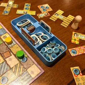 May include: A blue plastic container with compartments filled with blue buttons. The container is on a wooden table with a game board and game pieces. The game board is a patchwork quilt design with different colored squares. There are also several game cards with numbers and symbols on them.
