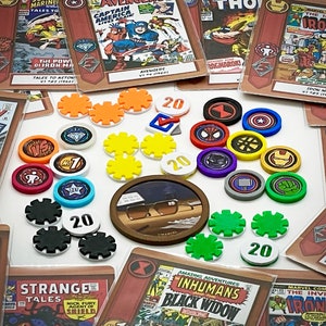 May include: A collection of colorful game pieces, including tokens with numbers, gears, and comic book characters. The pieces are arranged on a white surface with comic book covers in the background. The comic book covers feature characters like Captain America, Iron Man, and Black Widow.
