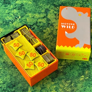 May include: An orange and yellow board game box with the text "MOVING WILD" and a hippo illustration. The game box is next to an open box with yellow game pieces and tokens with the number "50" on them. The game is on a green surface.