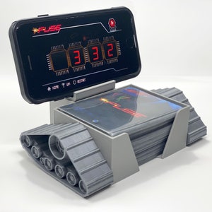 May include: A gray plastic tank-like base with a black smartphone on top. The phone screen displays a digital timer counting down from 3:32. The phone is attached to the base with a gray plastic stand. The base has a stack of gray cards inside.