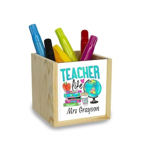 Puede incluir: Porta lápices de madera con un panel frontal blanco que presenta un gráfico colorido de un globo terráqueo, libros, una manzana y una taza de café. El texto "TEACHER life" está en turquesa con un corazón rosa. El texto "Mrs. Grayson" está debajo del gráfico.