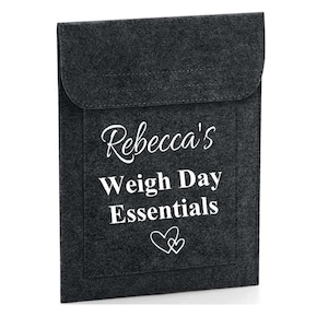 May include: A dark grey felt pouch with a flap closure. The front of the pouch features the text "Rebecca's Weigh Day Essentials" in white script and block letters, with two small heart outlines.