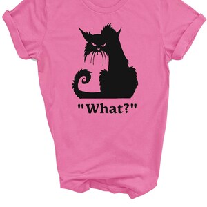 May include: Pink t-shirt featuring a black silhouette of a cat with an angry expression. The cat has pointed ears, a long tail, and a mustache. Below the cat is the word "What?" in black text.