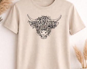 May include: A beige t-shirt featuring a black line drawing of a Highland cow's head. The illustration shows the cow's shaggy fur, horns, and facial features. The shirt is hanging on a wooden hanger.