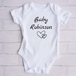 Może przedstawiać: Biały śpioszek dla niemowlęcia z czarnym napisem "Baby Robinson" i dwoma czarnymi sercami.
