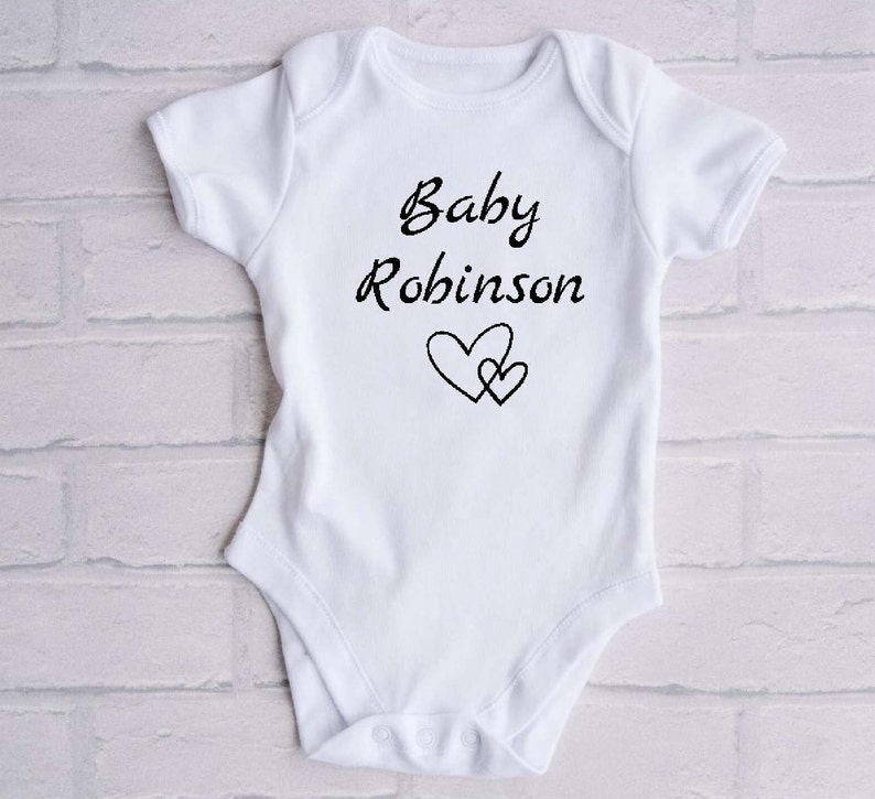 Może przedstawiać: Biały śpioszek dla niemowlęcia z czarnym napisem "Baby Robinson" i dwoma czarnymi sercami.