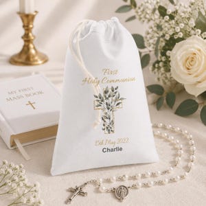 May include: White drawstring bag with "First Holy Communion" text, a cross design with leaves, and the date "15th May 2022". A rosary and a book with a cross are also visible.